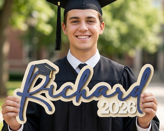 May include: A smiling graduate in a black cap and gown holds a wooden sign with the name "Richard" in navy blue script, a graduation cap graphic, and the year "2026". The sign has a cream-colored background and a navy blue outline.