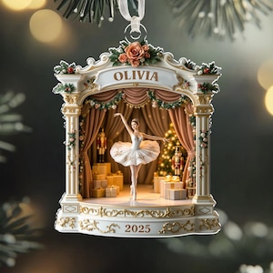 May include: A Christmas ornament featuring a ballerina on a stage. The ornament is white and gold, with the name "OLIVIA" above the stage and the year "2025" below. The ballerina is wearing a white tutu. The ornament is hanging from a white ribbon.