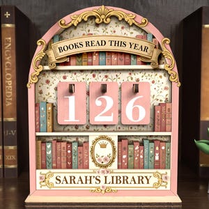 May include: A pink wooden bookshelf-shaped counter with the text "BOOKS READ THIS YEAR" and "SARAH'S LIBRARY". The counter displays the number "126" on pink panels. The bookshelf is decorated with floral patterns and miniature book illustrations.