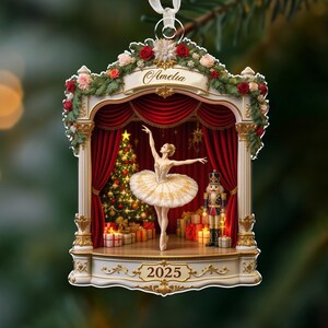 May include: A festive Christmas ornament depicting a ballerina on a stage, complete with a Christmas tree and a nutcracker. The ornament has a white ribbon for hanging and the name "Amelia". The year "2025" is displayed at the bottom.