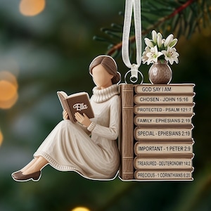 Könnte beinhalten: Weihnachtsschmuck mit einer Frau, die ein Buch mit dem Namen "Ella" liest. Die Frau sitzt neben einem Stapel Bücher mit religiösen Phrasen. Eine kleine Vase mit weißen Blumen steht auf den Büchern.