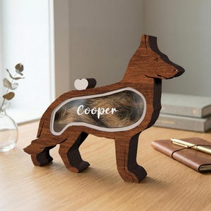 Peut inclure: Boîte souvenir en bois en forme de chien, avec une fenêtre transparente affichant de la fourrure et le nom "Cooper". La boîte est en bois brun foncé et repose sur quatre pattes. Un petit détail en forme de cœur blanc est à l'arrière.