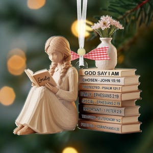 May include: A wooden ornament depicting a figure reading a book, with the name "Emily" on it. The ornament includes a stack of books with the text "GOD SAY I AM" and Bible verses. A small vase with flowers is also present.