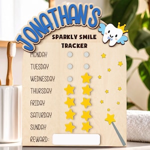 May include: Wooden "Jonathan's Sparkly Smile Tracker" with a tooth graphic and star stickers. The tracker has spaces for each day of the week and a reward section. Two toothbrushes in a white holder are on the left.