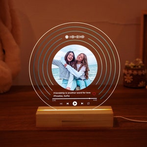 Może przedstawiać: Okrągła, przezroczysta lampa akrylowa z fotografią dwóch przyjaciółek na plaży w środku. Lampa ma drewnianą podstawę i światło, które świeci przez akryl. Na lampie widnieje tekst "Friendship is another word for love" wraz z imionami "Phoebe, Sofia".