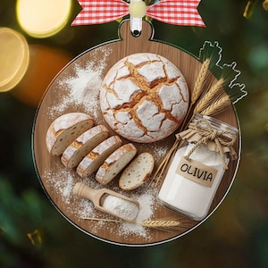 Op de afbeelding: Houten kerstornament in de vorm van een snijplank, met een brood, gesneden brood, bloem en een pot met het label "OLIVIA". Het ornament is versierd met een rood-witte geruite strik.