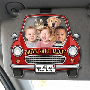 May include: A red car-shaped car visor clip featuring a photo of two children, a cat, and a dog. The text reads DRIVE SAFE DADDY and a license plate with LOVE Andy, Mia, Kelly, Ricoh, Tabby. The design includes headlights, a grille, and mirrors.