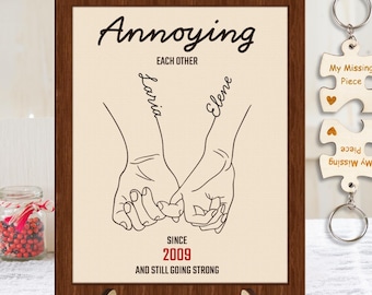 May include: A brown wooden frame with a white background and black text that reads "Annoying Each Other" with two names written in cursive, "Laria" and "Elene", and a line drawing of two hands holding each other's pinky fingers. The text "Since 2009 And Still Going Strong" is below the drawing. A wooden keychain with two puzzle pieces that read "My Missing Piece" is on the right side of the frame.