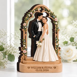 May include: A wooden wedding cake topper featuring a couple standing under a decorative arch adorned with small pink and blue flowers. The base is engraved with the text WILLIAM & OLIVIA and the date 05.02.2026, flanked by two small heart icons.