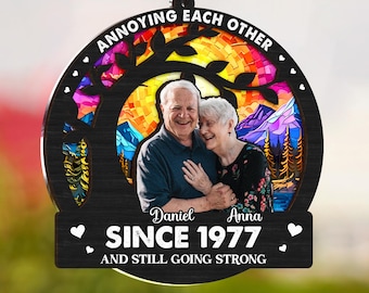 May include: A circular wooden ornament featuring a photograph of an elderly couple. The text "ANNOYING EACH OTHER" arcs across the top, with "SINCE 1977 AND STILL GOING STRONG" below. The ornament is suspended by a black chain.