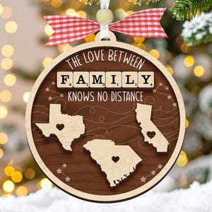 Può includere: Ornamento natalizio in legno con la scritta "THE LOVE BETWEEN FAMILY KNOWS NO DISTANCE". L'ornamento presenta tre ritagli di stato con dettagli a cuore, un fiocco a scacchi rosso e bianco e un nastro bianco per appenderlo.