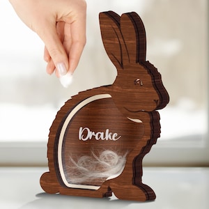 May include: A wooden rabbit figurine with a cutout window, the name "Drake" is engraved on the window, and white fur is inside the window.