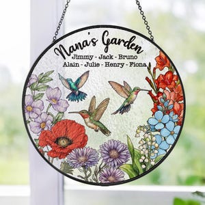 May include: A round stained glass window hanging with a black metal frame. The window features a floral design with hummingbirds and the text "Nana's Garden" with a list of names: Jimmy, Jack, Bruno, Alain, Julie, Henry, Fiona.
