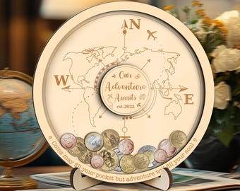 May include: A wooden coin bank with a world map design. It features a compass rose, an airplane, and the text "Our Adventure Awaits est.2023." Coins are visible at the bottom. The text "Coins may fill your pocket but adventure will fill your soul" is also present.