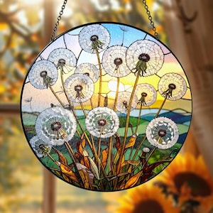 Puede incluir: Arte de ventana de vidriera redonda con un diseño de diente de león. La obra de arte muestra cabezas de semillas de diente de león blancas sobre un fondo de un paisaje colorido con un cielo azul y un sol amarillo. La pieza está enmarcada en negro y cuelga de una cadena.
