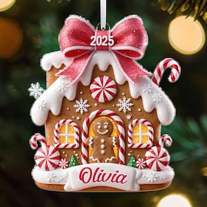 May include: Gingerbread house Christmas ornament with a red bow and the year 2025. The ornament features white icing, candy canes, and the name "Olivia" on a banner. The gingerbread man is in the center.