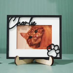 May include: Black wooden picture frame with a white mat and a photo of a ginger cat. The frame has a paw print cutout and the name "Charlie" written in cursive on the top.