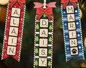 Puede incluir: Tres adornos navideños personalizados con nombres escritos con fichas de Scrabble. Cada adorno tiene una cinta, un lazo rojo y un diseño decorativo. Los nombres son Alain, Daisy y Marin.