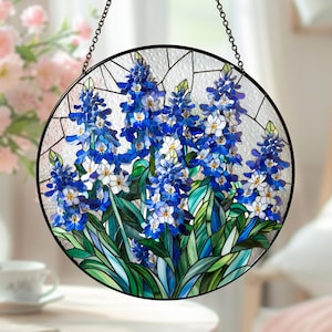 以下が含まれることがあります： 青と白の花と緑の葉が描かれた丸いステンドグラスパネル。 花柄のデザインは、透明で質感のある背景に配置され、黒い金属の縁と吊り下げチェーンで縁取られています。 装飾的なアートワークです。