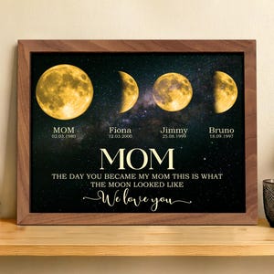 Op de afbeelding: Een ingelijste print met vier maanfasen tegen een sterrenachtergrond. De print bevat het woord "MOM" en de tekst "THE DAY YOU BECAME MY MOM THIS IS WHAT THE MOON LOOKED LIKE". Namen en data zijn ook inbegrepen.