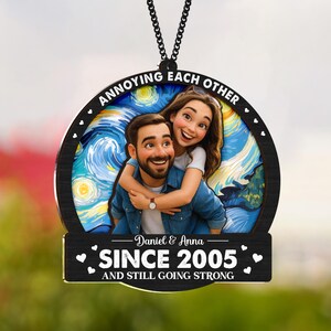May include: A round, black wooden ornament with a couple embracing, set against a starry night background. The text "ANNOYING EACH OTHER" arcs across the top. Below, it reads "Daniel & Anna SINCE 2005 AND STILL GOING STRONG."