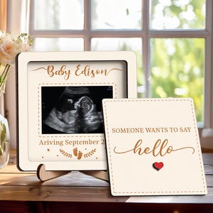 May include: A cream-colored wooden frame with a baby ultrasound image, the name "Baby Edison", and the text "Arriving September 2". A small card next to it says "Someone wants to say hello" with a red heart.