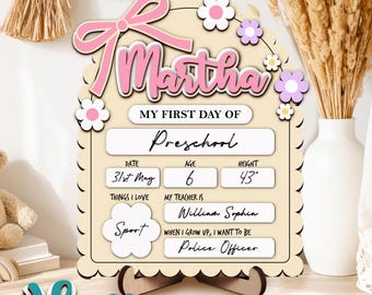 May include: A wooden sign for a child's first day of preschool, featuring the name "Martha" in pink. The sign includes details like date, age, height (43 inches), and interests. A separate teal name "Henry" is also present.