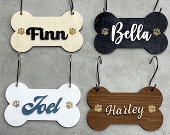 Könnte beinhalten: Vier hölzerne Knochen-Anhänger mit personalisierten Namen, "Finn", "Bella", "Joel" und "Harley", jeweils mit einem goldenen Pfotenabdruck auf jeder Seite.