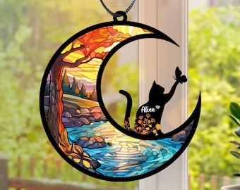 Può includere: Un acchiappasole in vetro colorato a forma di mezzaluna con una silhouette di gatto seduta sulla luna. Il gatto sta raggiungendo una farfalla. La luna presenta un design in vetro colorato con una scena di tramonto. Il nome del gatto, "Alice", è scritto sulla luna.