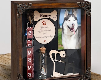 May include: A wooden shadow box memorial for a pet. It features a photo of a husky dog, a dog collar with a bell, a small glass vial with a rainbow charm, a wooden dog silhouette, and a bone-shaped plaque with the name "Henry" and the dates "2017" and "2023". The plaque also has the text "You were my favourite hello and my hardest goodbye."