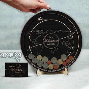 May include: A black wooden coin bank shaped like a world map with a plane flying around it. The text "Our Adventure Awaits" is written on the bank. A small black tag with the text "And so... the Adventure Begins" is in the lower left corner of the image.