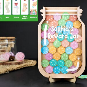 Könnte beinhalten: Ein Holz-Belohnungs-Glas mit einer durchsichtigen Vorderseite und einem abnehmbaren Deckel. Das Glas ist mit bunten Smiley-Gesichtern und dem Text "Sophia's Reward Jar" verziert.