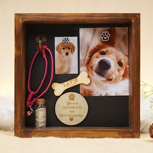 May include: A wooden shadow box memorial for a pet, featuring a pink collar, a small bottle, a bone-shaped wooden plaque with the text "La Vie 17.09.2022", a photo of a golden retriever puppy, and a wooden circle with the text "You were my favourite hello and my hardest goodbye".
