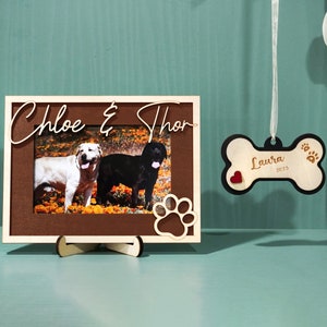 May include: A wooden picture frame with a brown background and a photo of two dogs, one white and one black, with the text "Chloe & Thor" above the photo. A wooden bone-shaped ornament with the text "Laura 2023" and a red heart is hanging from a white ribbon.
