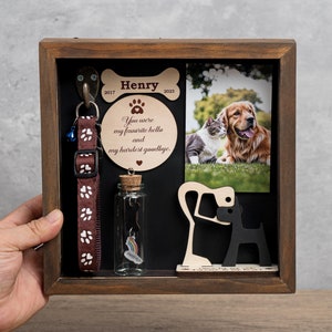 May include: A wooden shadow box memorial for a pet named Henry. The box includes a photo of a golden retriever and a cat, a dog collar, a small glass bottle with a rainbow charm, a wooden paw print, and a plaque with the text "You were my favorite hello and my hardest goodbye."