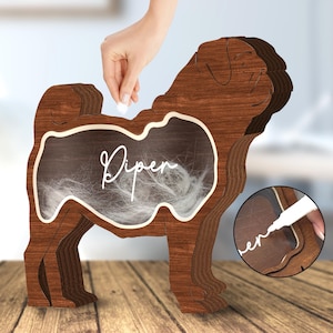 May include: A wooden pug-shaped pet hair catcher with a chalkboard insert. The chalkboard is labeled "Piper" and has white pet hair inside. A hand is shown adding more pet hair to the catcher.