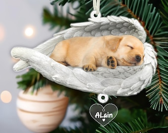 May include: White ceramic Christmas ornament featuring a sleeping golden retriever puppy nestled in a pair of angel wings. A heart-shaped tag with the name "ALain" hangs from the ornament.
