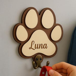 May include: A wooden paw print wall hook with the name "Luna" engraved. It holds a red leather dog collar with a silver buckle. The paw print has a brown border and cream-coloured pads. The hook is attached to a wall.