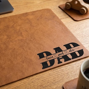 May include: A brown leather desk mat with the word "DAD" in black, with "laura ♡ jacob ♡ alex" underneath. The mat is rectangular and has a textured surface. A laptop and a wooden toy car are also visible.