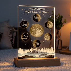 May include: A lit acrylic sign with a moon and stars design. The sign reads "We love you to the moon and back". There are moons with names and dates on them. The sign is on a wooden base.