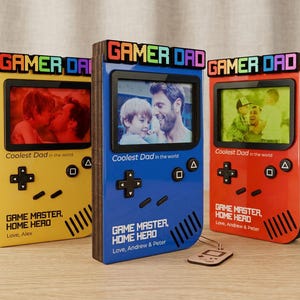 Op de afbeelding: Drie kleurrijke fotolijsten in de vorm van retro gameconsoles. Elke lijst heeft een andere kleur: geel, blauw en rood. De lijsten hebben de woorden "GAMER DAD" bovenaan en "Coolest Dad in the world" onderaan. Elke lijst bevat een foto.