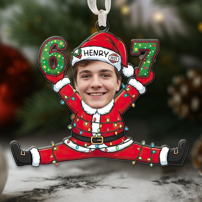 May include: A personalized Christmas ornament featuring a smiling face in a Santa Claus suit, holding the numbers 6 and 7. The ornament has the name "HENRY" and the year "2025" on the Santa hat. The ornament is red, green, and white.