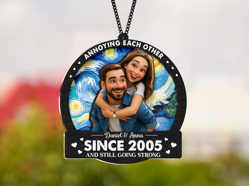 May include: A round, black wooden ornament with a couple embracing, set against a starry night background. The text "ANNOYING EACH OTHER" arcs across the top. Below, it reads "Daniel & Anna SINCE 2005 AND STILL GOING STRONG."