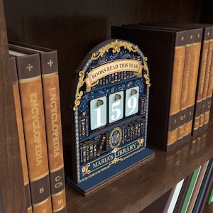 May include: A wooden book counter with a navy blue background and gold accents. The counter reads "BOOKS READ THIS YEAR" and displays the number "159". The bottom of the counter reads "MARIA'S LIBRARY". The counter is on a wooden shelf with books.