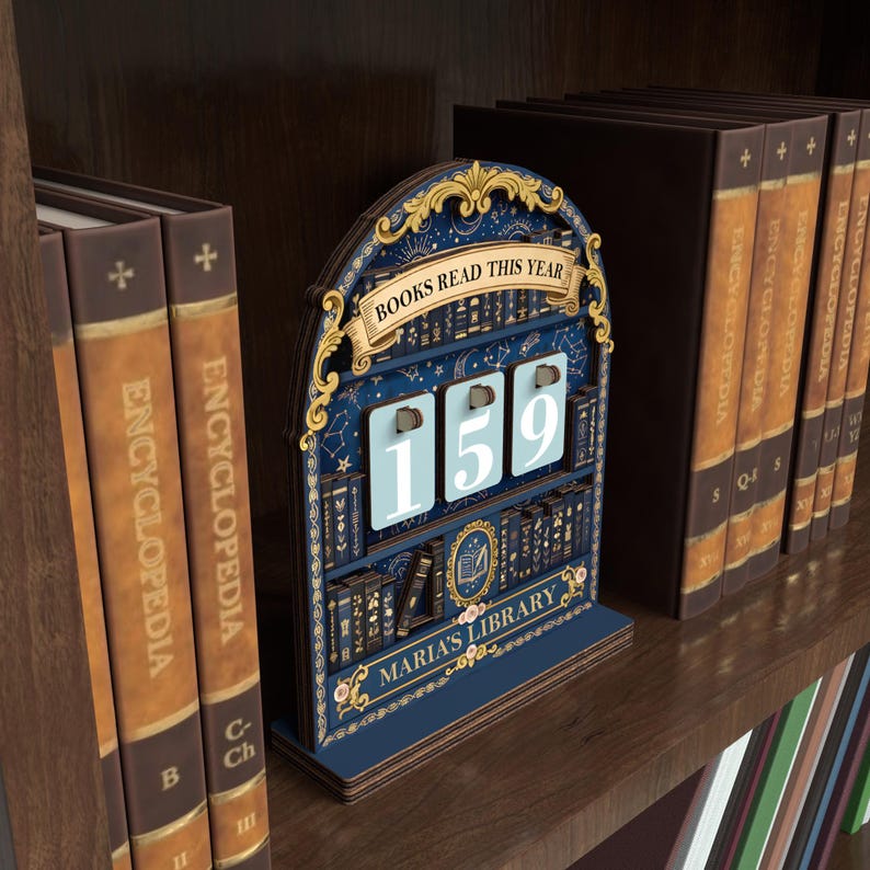 May include: A wooden book counter with a navy blue background and gold accents. The counter reads "BOOKS READ THIS YEAR" and displays the number "159". The bottom of the counter reads "MARIA'S LIBRARY". The counter is on a wooden shelf with books.