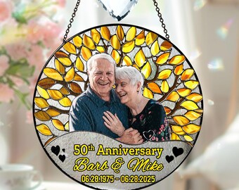 May include: A round, stained-glass window with a photo of an elderly couple, celebrating their 50th anniversary. The window features a yellow and brown leaf design, with the text "50th Anniversary Barb & Mike 06.28.1975-06.28.2025". A heart-shaped crystal hangs above.