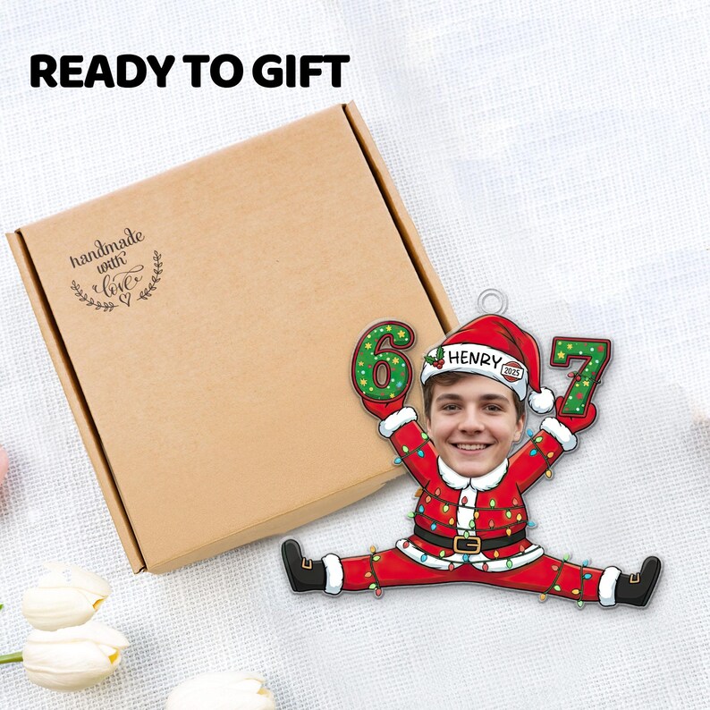 May include: A personalized Christmas ornament featuring a person's face in a Santa Claus suit, holding the numbers 6 and 7. The ornament is decorated with Christmas lights and the name "Henry" is visible. The words "READY TO GIFT" are at the top.