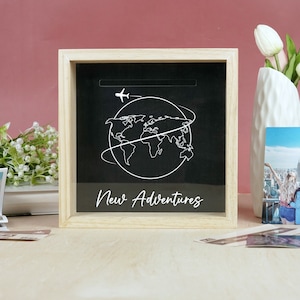 May include: A wooden shadow box with a black background and a white outline of a world map with an airplane above it. The text "New Adventures" is written below the map.