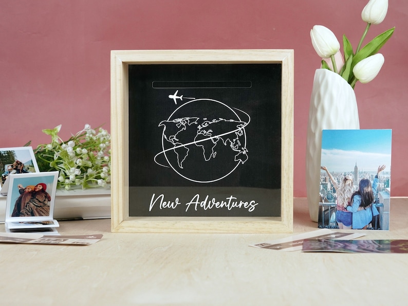 May include: A wooden shadow box with a black background and a white outline of a world map with an airplane above it. The text "New Adventures" is written below the map.