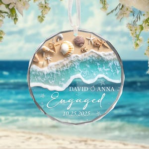 May include: A round, clear glass ornament with a beach scene. The ornament features sand, seashells, and turquoise water with the word "Engaged" and the names "David & Anna" and the date "10.25.2025". A white ribbon is attached for hanging.
